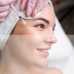 Close up of hands of young cosmetologist injecting botox in female face. She is standing and smiling. The woman is closed her eyes with relaxation.